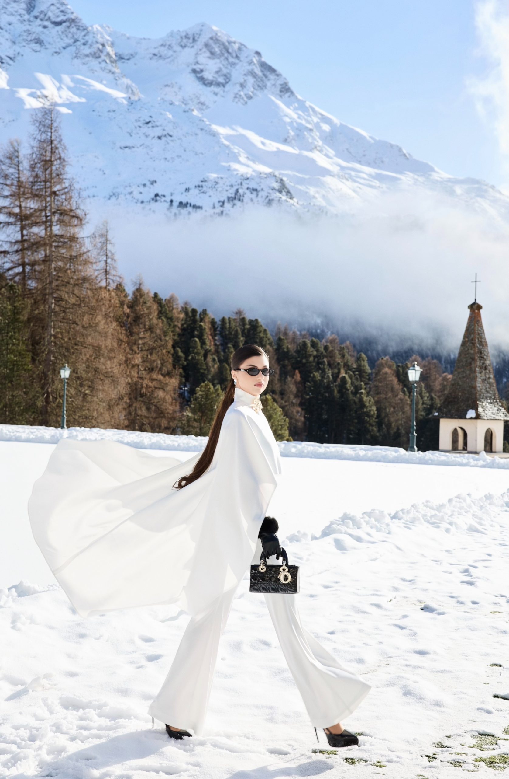 Full-length luxury fashion editorial, model walking in white cape and wide-leg trousers with black Lady Dior bag and fur gloves, St. Moritz Alpine landscape, photography by Furkan Ramadani