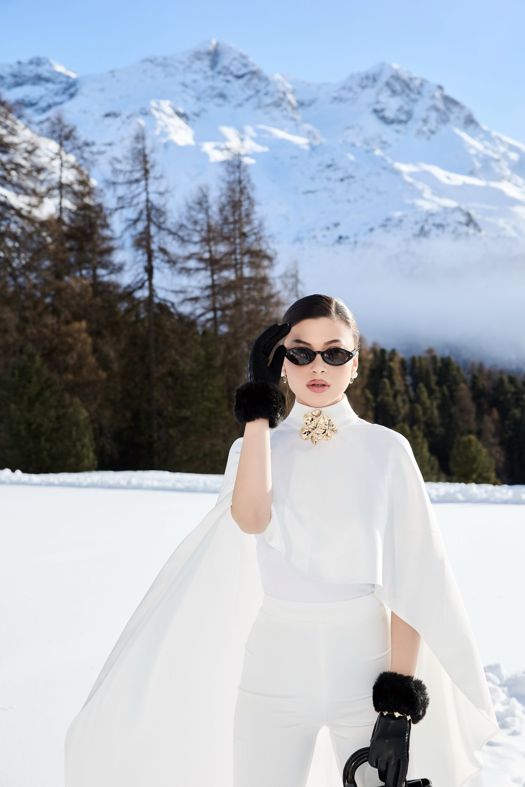 Luxury fashion editorial in St. Moritz, model in white cape ensemble with gold brooch and black fur gloves against snow-covered Alpine peaks, photography by Furkan Ramadani