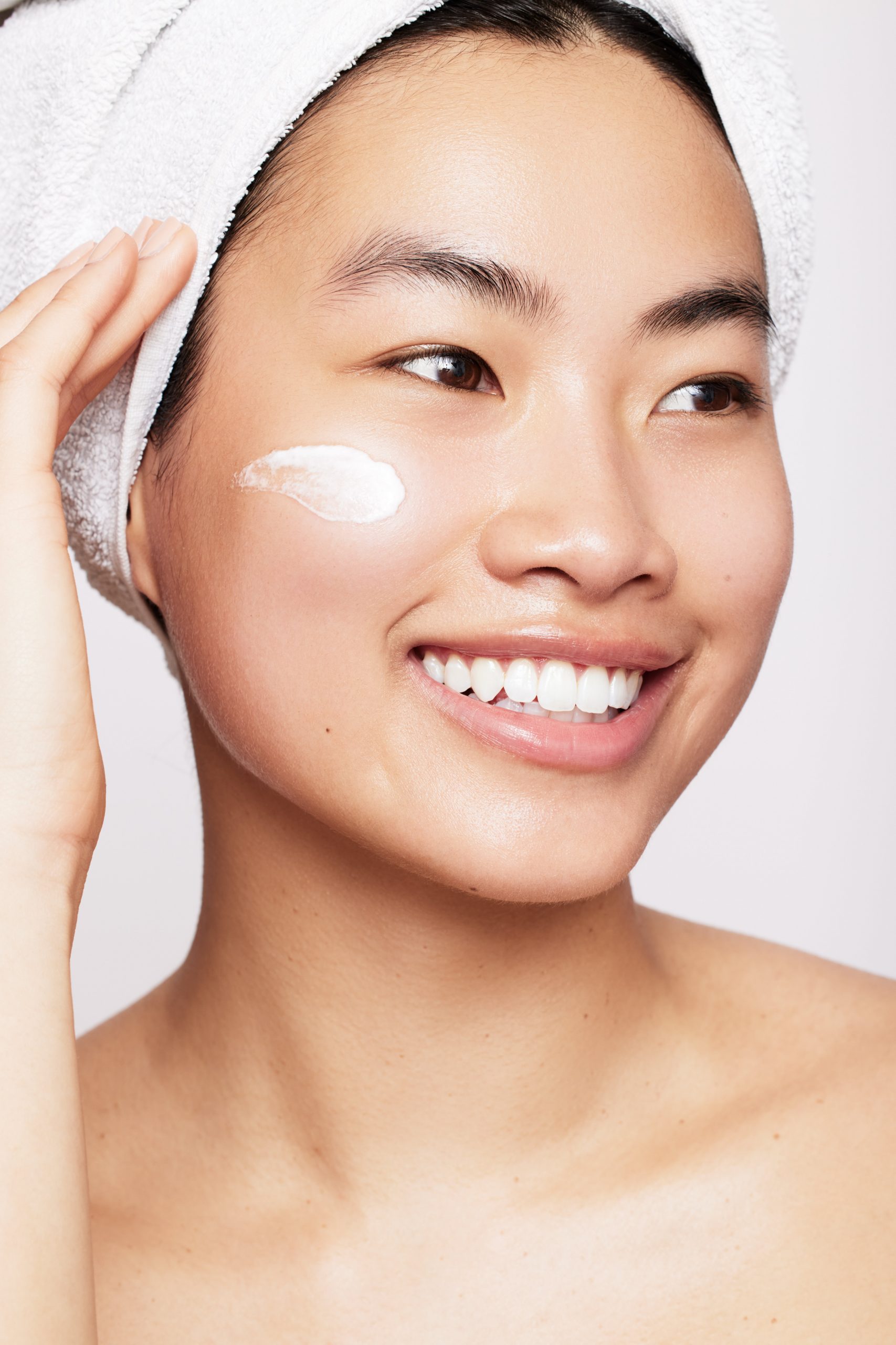 Skincare beauty portrait, smiling model with cream swatch on cheek and towel wrap, commercial skincare photography by Furkan Ramadani