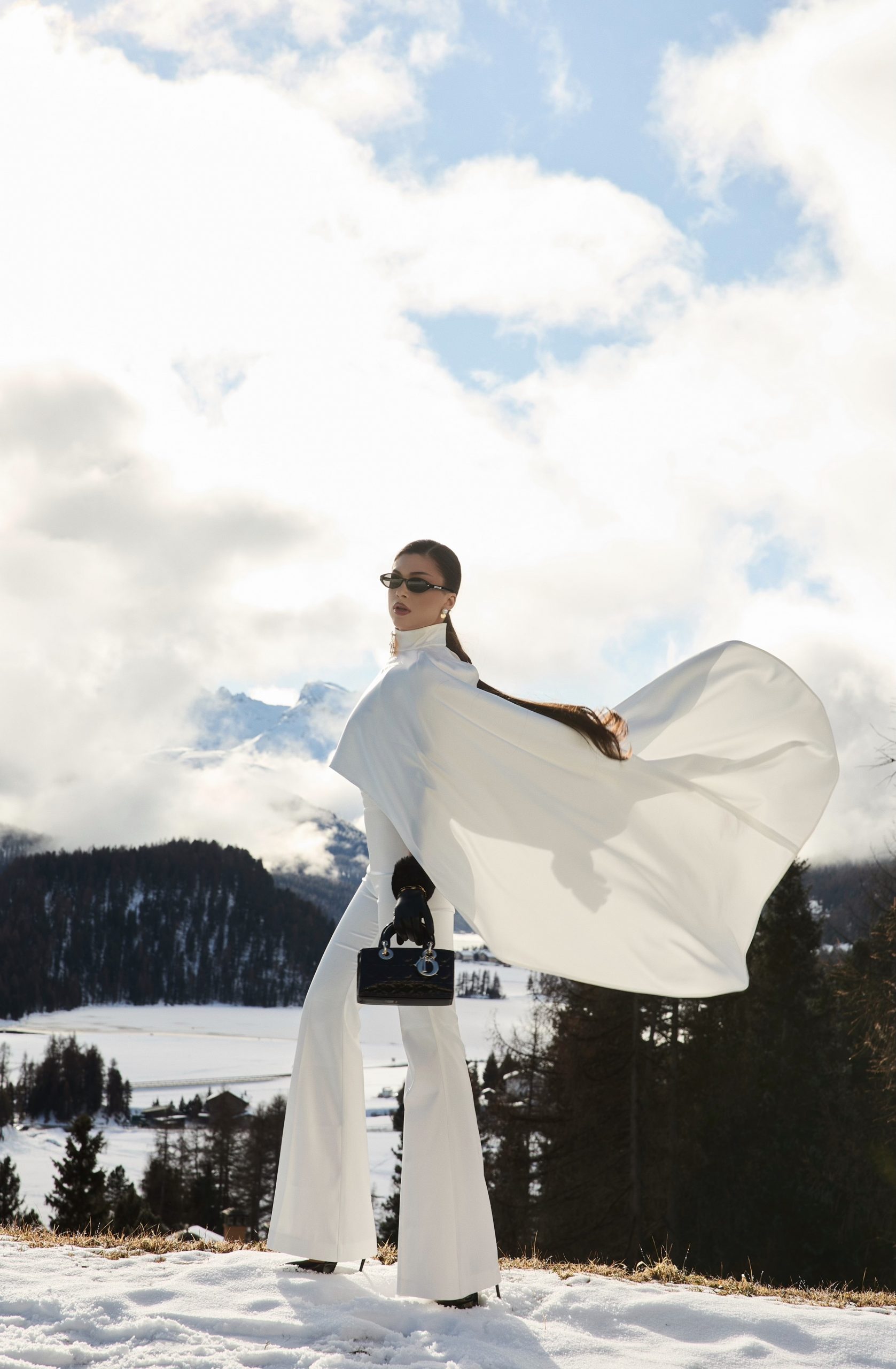 Dramatic fashion editorial, model in flowing white cape against stormy Alpine sky, fabric sweeping in wind with snowy mountain valley below, St. Moritz, photography by Furkan Ramadani